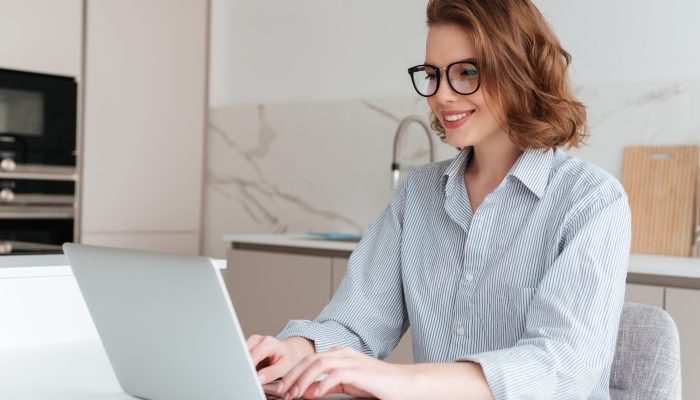 Elegant smiling woman in glasses and striped shirt using laptop computer while siting at table in kitchen